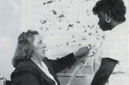 Two women engage in a discussion; one points to a map on the wall, while the other listens attentively, holding a paper about local home care services.
