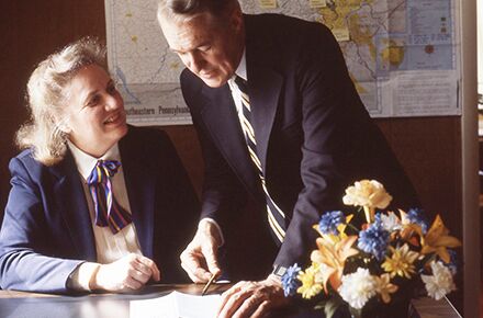 Two people in business attire are discussing a document at a desk with a bouquet of flowers on it. A map hangs on the wall, hinting at their discussion possibly involving local home care services or elderly care assistance.