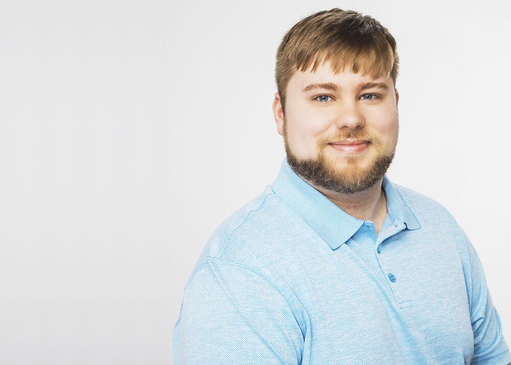 A young man with a beard and short hair wearing a light blue polo shirt smiles at the camera, showcasing his expertise in caregiving services.