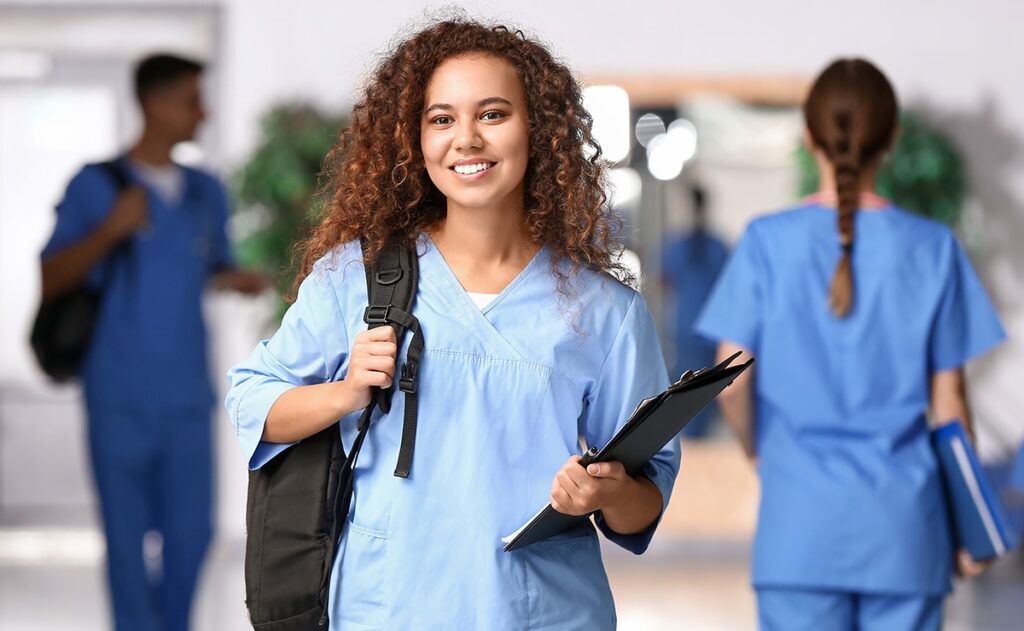 A young female healthcare professional in blue scrubs, holding a clipboard and backpack, smiles at the camera. She represents local home care services, ensuring personalized attention for those in need.