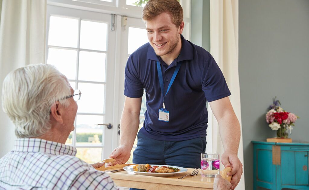 A caregiver from local home care services serves a meal to an elderly person sitting at a table in a well-lit room.