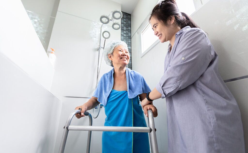 An elderly woman using a walker stands in a shower area with the assistance of a younger woman, both smiling. This scene highlights the importance of personal care services in providing elderly care assistance.
