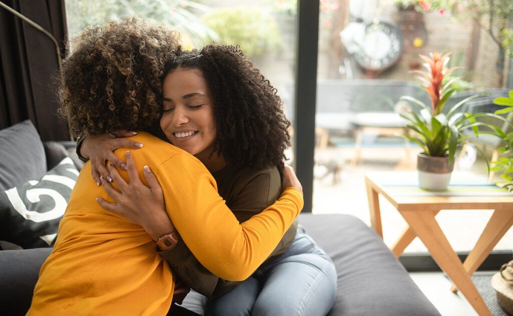 Two people sit on a couch in a warm embrace, one wearing a yellow top and the other in a green top, with a plant nearby.