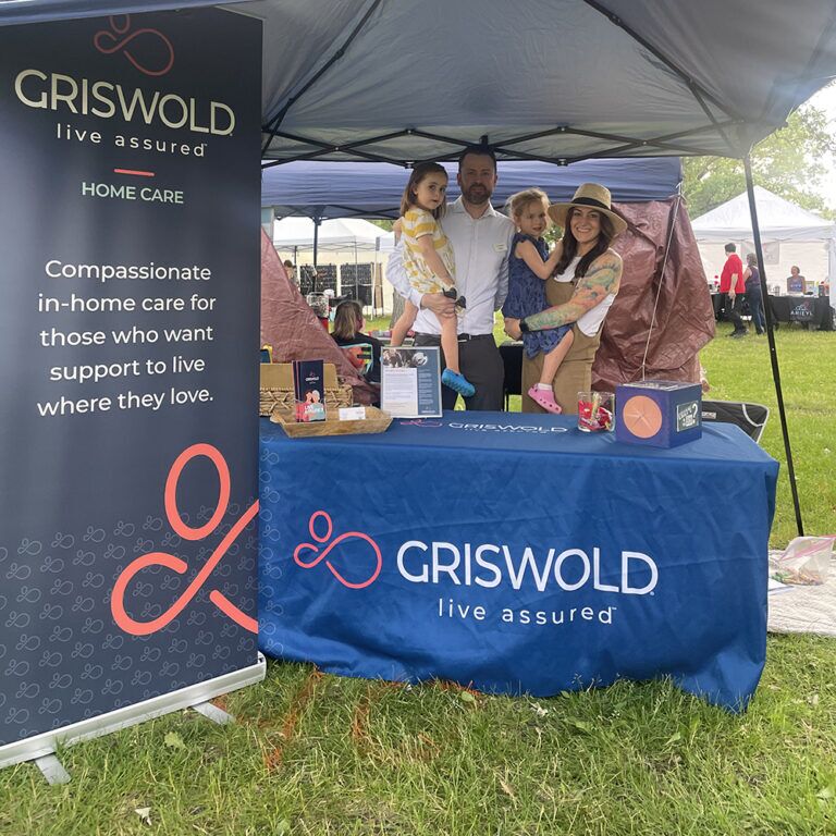 A family posing under a canopy beside a Griswold Home Care booth at an outdoor event, showcasing their caregiving services.