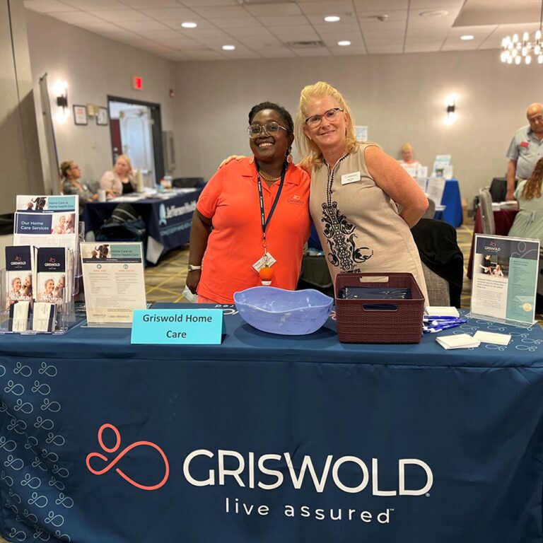 Two women stand smiling behind a table set up with brochures and a large sign reading "Griswold Home Care" at an event, promoting elderly care assistance.