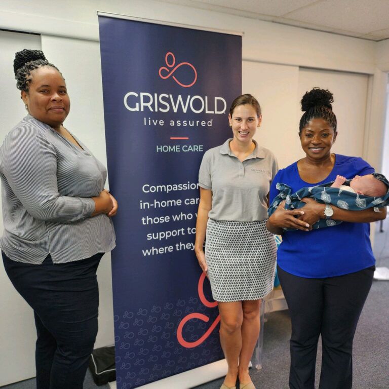 Three women stand in front of a Griswold Home Care banner; one woman is holding a baby, showcasing their commitment to local home care services.