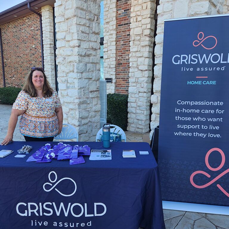 A woman stands behind a table covered with promotional materials and a blue tablecloth branded with "Griswold - Live Assured" next to a banner for Griswold Home Care, highlighting their in-home care services.