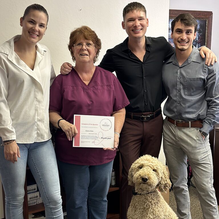 Four people standing indoors, one holding a certificate related to local home care services. A dog sits at their feet.