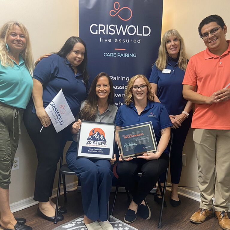 Six individuals pose for a group photo in front of a Griswold Care Pairing banner. Two seated women hold framed recognitions, one showcasing "20 Steps" and the other "Great Place to Work." Their dedication to in-home care services shines through these awards.