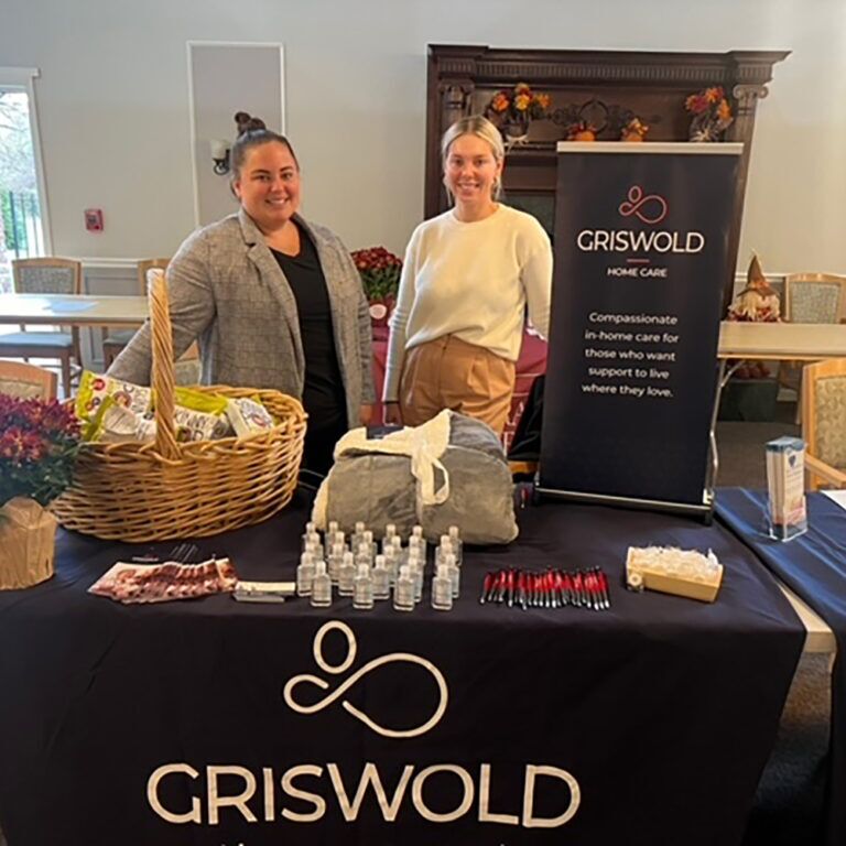 Two women stand behind a table with a Griswold Home Care banner, displaying informational materials, pens, hand sanitizers, and a gift basket. The setting appears to be a community event highlighting in-home care services.