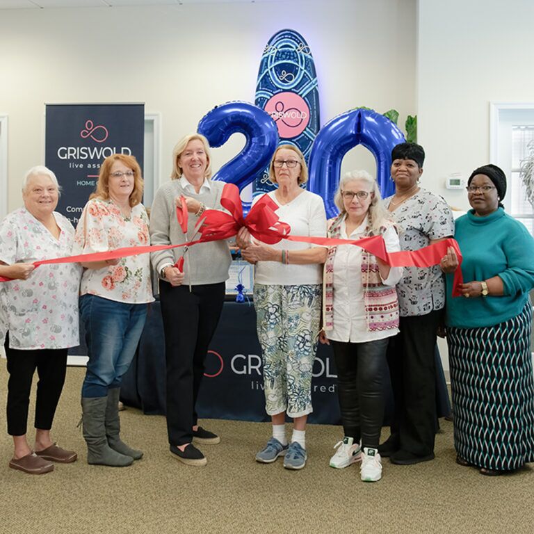 A group of seven individuals stand together holding a large red ribbon and scissors, celebrating Griswold's 20th anniversary with a sign and balloons, symbolizing their commitment to providing exceptional caregiver services.