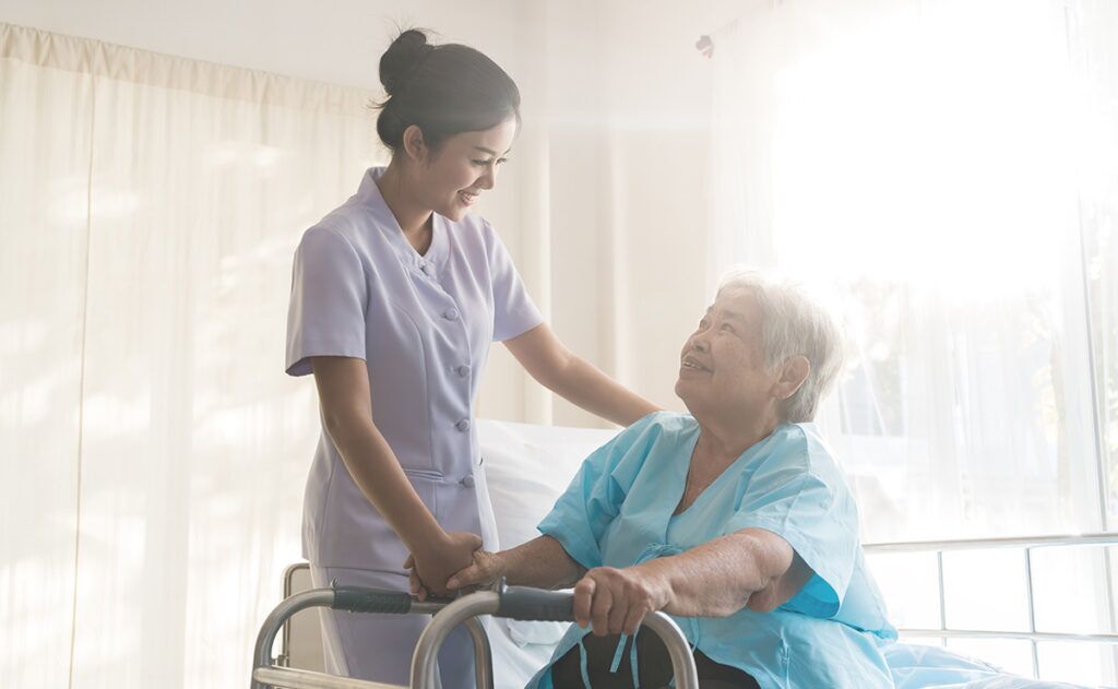 A caregiver provides elderly care assistance and smiles at an elderly person using a walker in a sunlit room.
