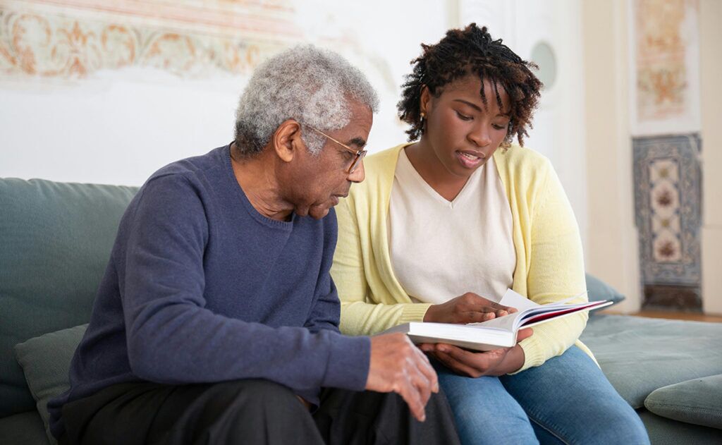 An elderly man and a young woman sit on a couch, focusing on a book the woman is holding open. The man looks at the book intently while the woman gestures to a page, exemplifying attentive in-home care services.