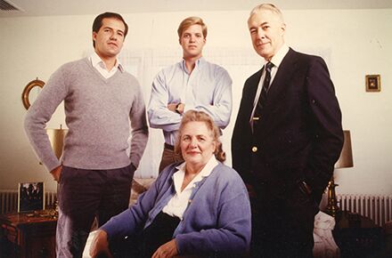 A group photo features three men standing and one woman seated in a living room. Two men wear sweaters, one wears a suit, and the woman wears a sweater over a blouse. The group is discussing personal care services.