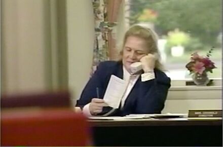 A woman in a dark blazer sits at a desk, holding a phone to her ear and looking at a piece of paper. A nameplate and a flower vase are on the desk, suggesting she might be coordinating elderly care assistance.