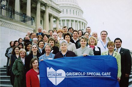 A large group of people stands together on the steps of a government building, holding a blue banner that reads "GRISWOLD SPECIAL CARE." The U.S. Capitol building is visible. They are advocating for local home care services.