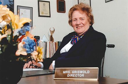 A woman in formal attire is seated at a desk with a nameplate reading "Mrs. Griswold, Director." Various certificates and a trophy are displayed on the wall. Flowers are in the foreground, highlighting her dedication to caregiving services.