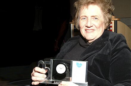 An elderly woman with short hair, dressed in black, smiles while holding a framed circular item with a "25" on one side and a blue heart logo on the other, representing her appreciation for exceptional caregiving services.