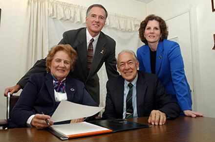 Four individuals, three seated and one standing, pose for a photo in an office-like setting. Two women and two men, dressed in business attire, appear around a desk with papers discussing caregiver services.