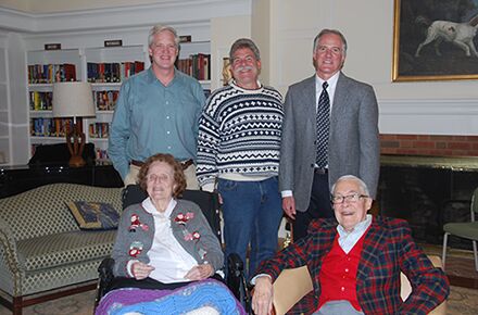 A group of people posing for a photo indoors; three men are standing behind two seated elderly individuals, showcasing the warmth and comfort typical of in-home care services.