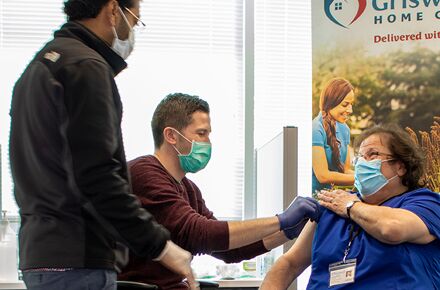 A healthcare professional administers a vaccine to a seated woman while another person stands nearby. All individuals are wearing masks, highlighting the importance of personal care services in preventive health measures.
