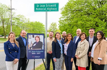 A group of people stands beside a sign reading "Dr. Jean Griswold Memorial Highway," with a commemorative poster of Dr. Jean Griswold placed below the sign, honoring her contributions to local home care services.