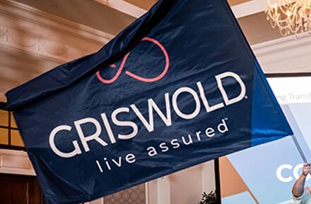 A blue flag with "GRISWOLD live assured" written on it is displayed indoors, promoting their local home care services.