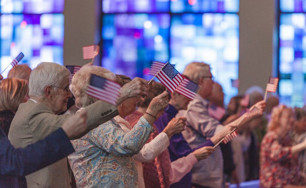 A group of people, many elderly, hold small American flags while standing in front of large stained glass windows. Among them are professionals offering elderly care assistance, ensuring everyone is comfortable and happy.