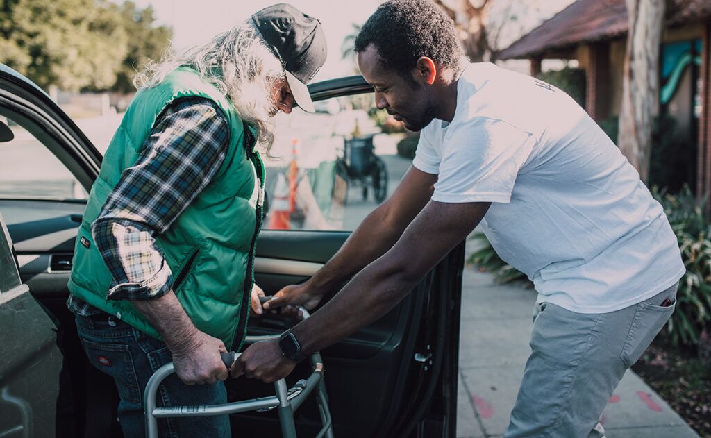 A man with a walker is assisted by another man to enter a car, highlighting the importance of caregiving services. The surroundings include trees, a sidewalk, and a building.