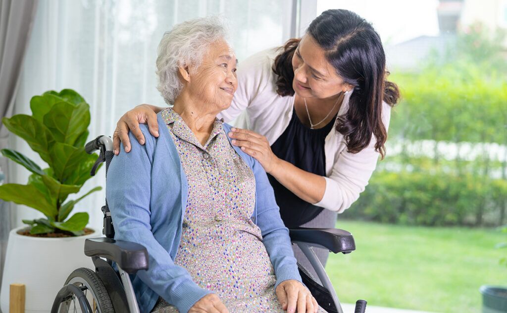An elderly woman in a wheelchair is smiling at a younger woman who is standing beside her, gently touching her shoulders. They are indoors with a window showing greenery. This serene moment highlights the importance of caregiver services and the joy they bring to daily life.