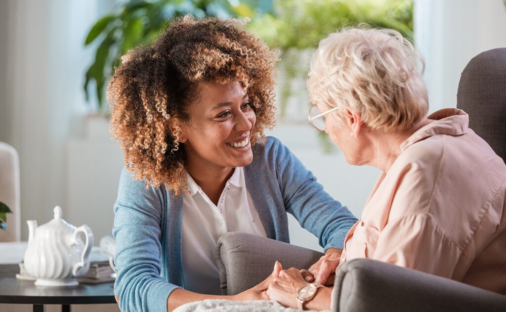 A young woman with curly hair smiles while conversing with an older woman sitting in an armchair. Providing elderly care assistance, she ensures the older woman's comfort as a white teapot sits on a nearby table.