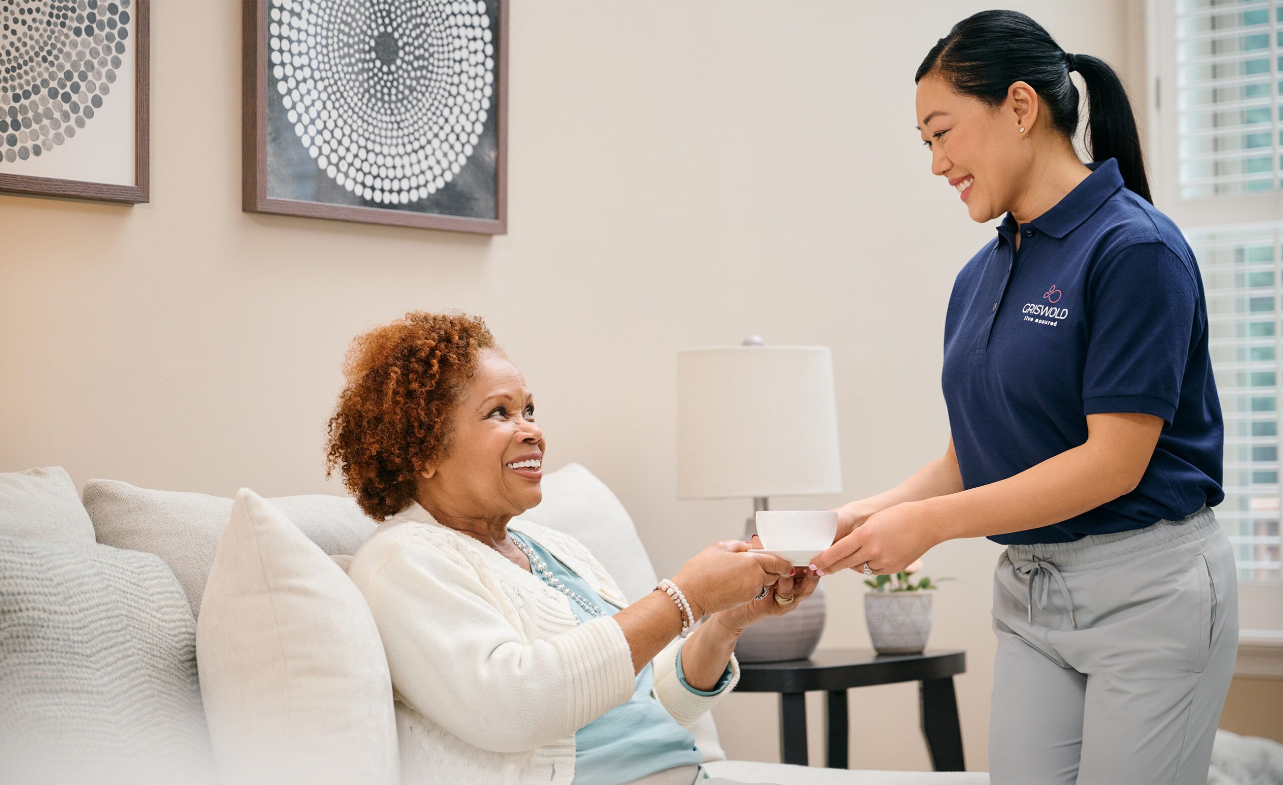 A caregiver from a home health care service, dressed in a blue uniform, hands a smiling elderly woman a small object while they sit on a beige sofa in the well-lit living room.