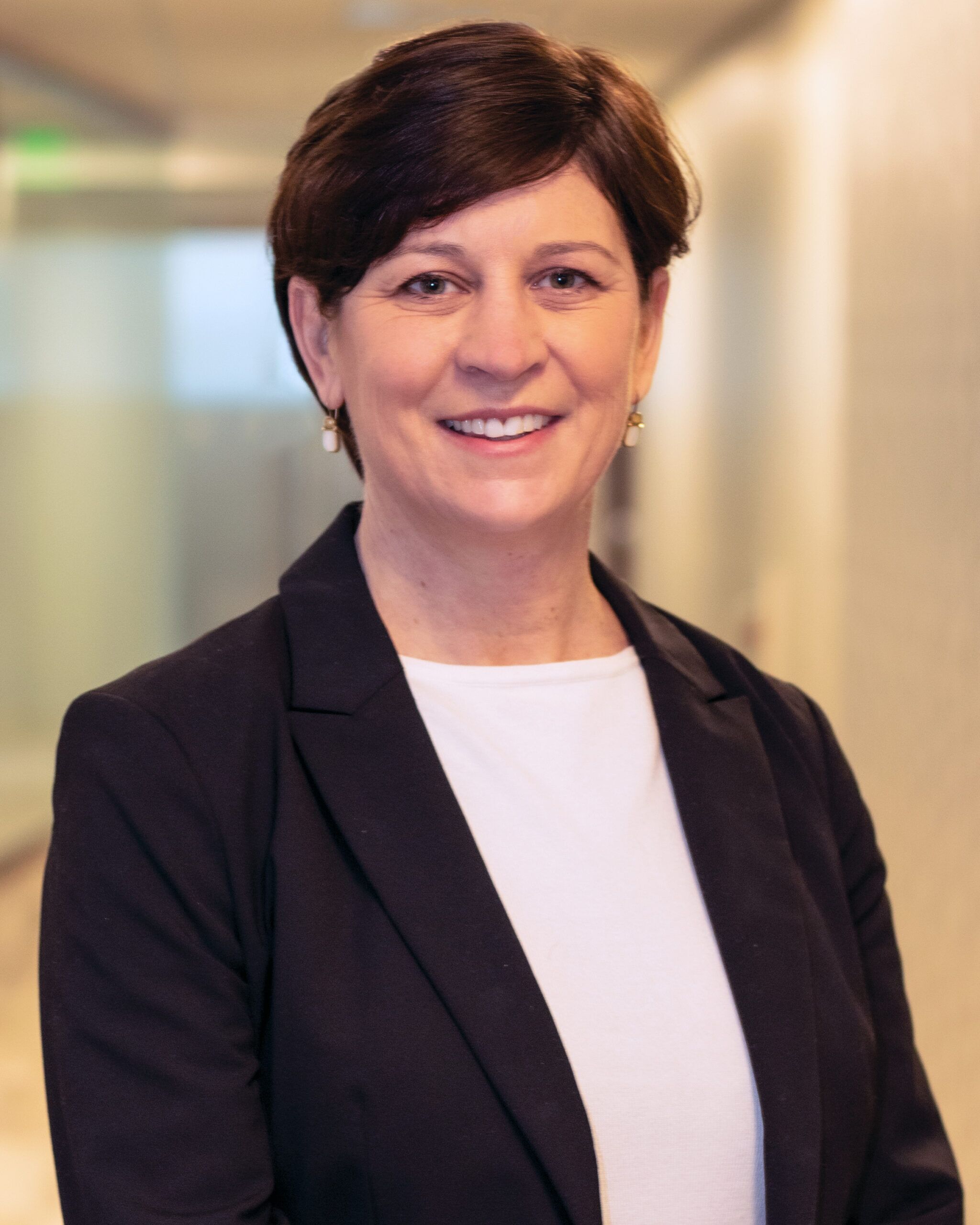 A person with short brown hair, wearing a black blazer and white shirt, smiles while standing in a corridor with frosted glass walls, reflecting their dedication to elder home care services.