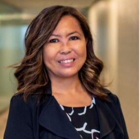 A woman with shoulder-length wavy hair smiles while standing in a well-lit indoor setting, wearing a dark blazer over a patterned blouse, radiating the dedication and compassion often seen in elder home care services.