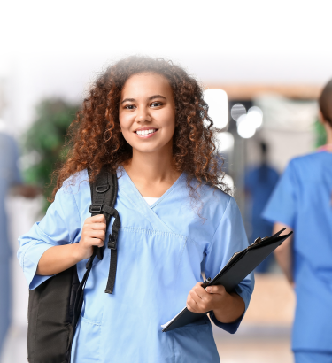 A smiling nurse holds a clipboard with her left hand and carries a backpack over her right shoulder. She is standing in a medical facility, wearing blue scrubs. Other healthcare professionals are in the background, dedicated to providing exceptional personal care services.