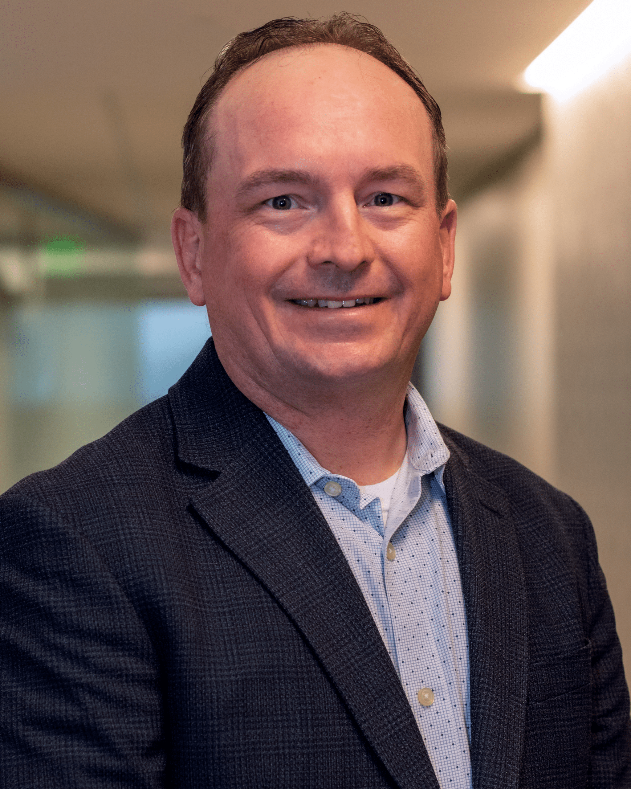 A man wearing a dark blazer and light blue button-up shirt stands smiling in a hallway with blurred background, embodying the professionalism often associated with personal care services.