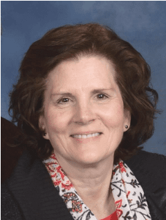 A woman with short, wavy brown hair, wearing a black sweater and a patterned scarf, smiles at the camera against a blue background, embodying the warmth and compassion of our personal care services.