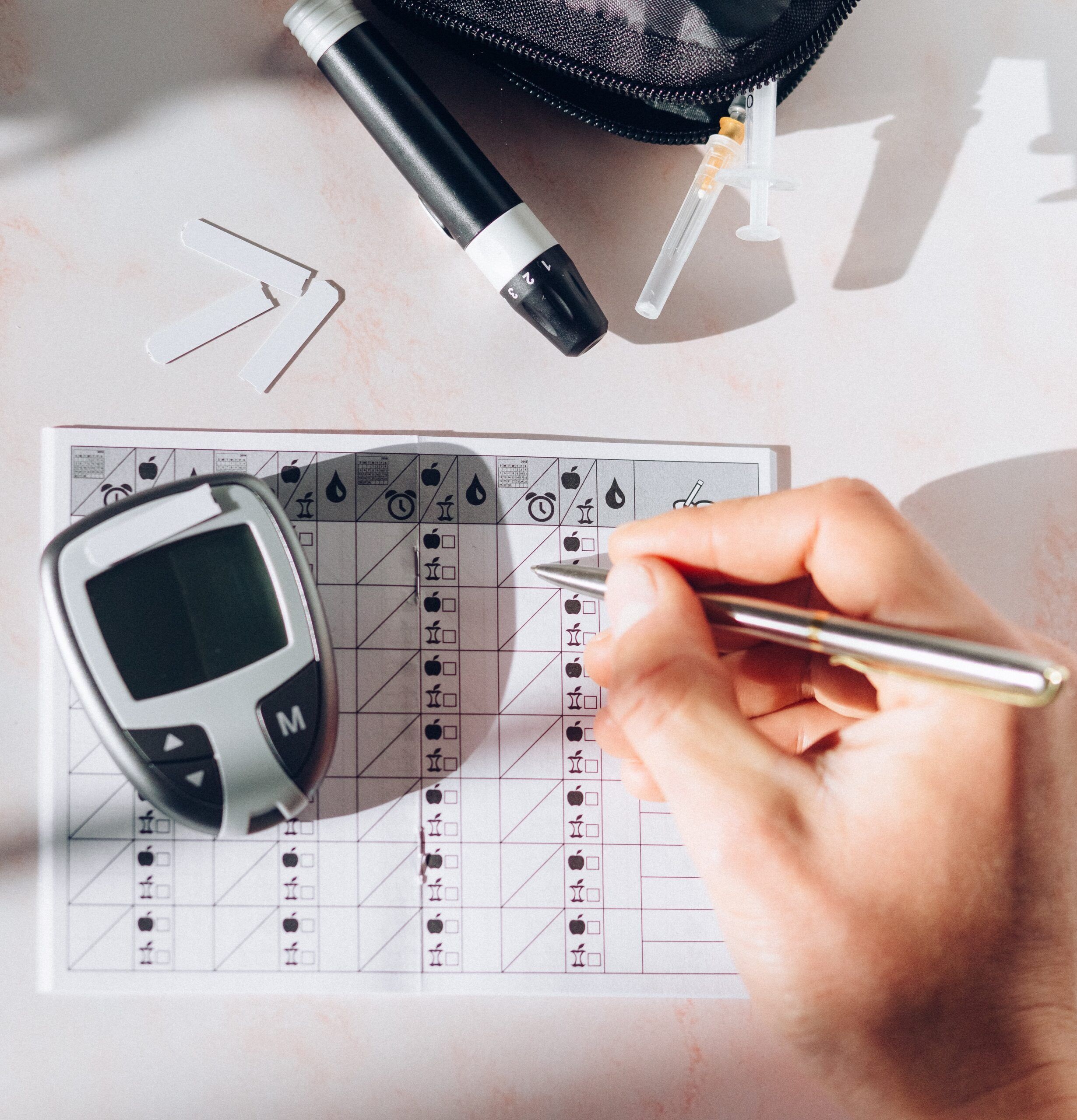 A hand holding a pen writes on a diabetes tracking sheet with a blood glucose meter