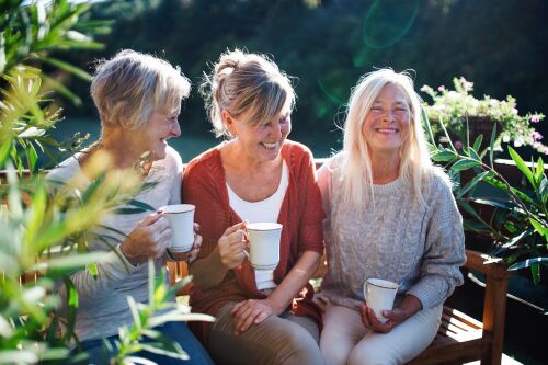 Three women sitting on a bench outdoors, smiling and holding coffee mugs