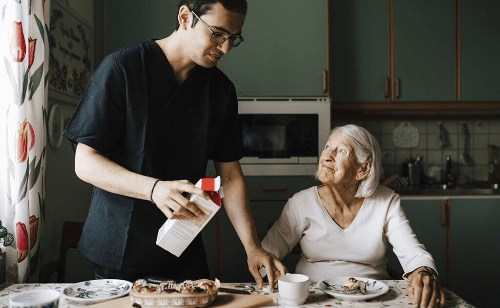 A caregiver in scrubs pours milk into a mug for an elderly woman